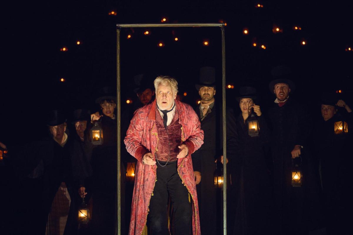 Erik Thomson as Scrooge. He is wearing a pink coat and is standing in a door frame. The ensemble are in black behind him.