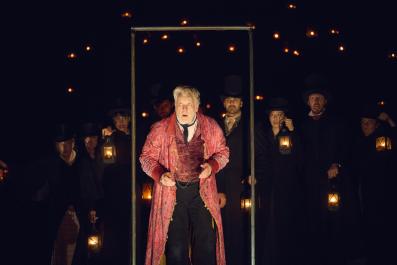 Erik Thomson as Scrooge. He is wearing a pink coat and is standing in a door frame. The ensemble are in black behind him.