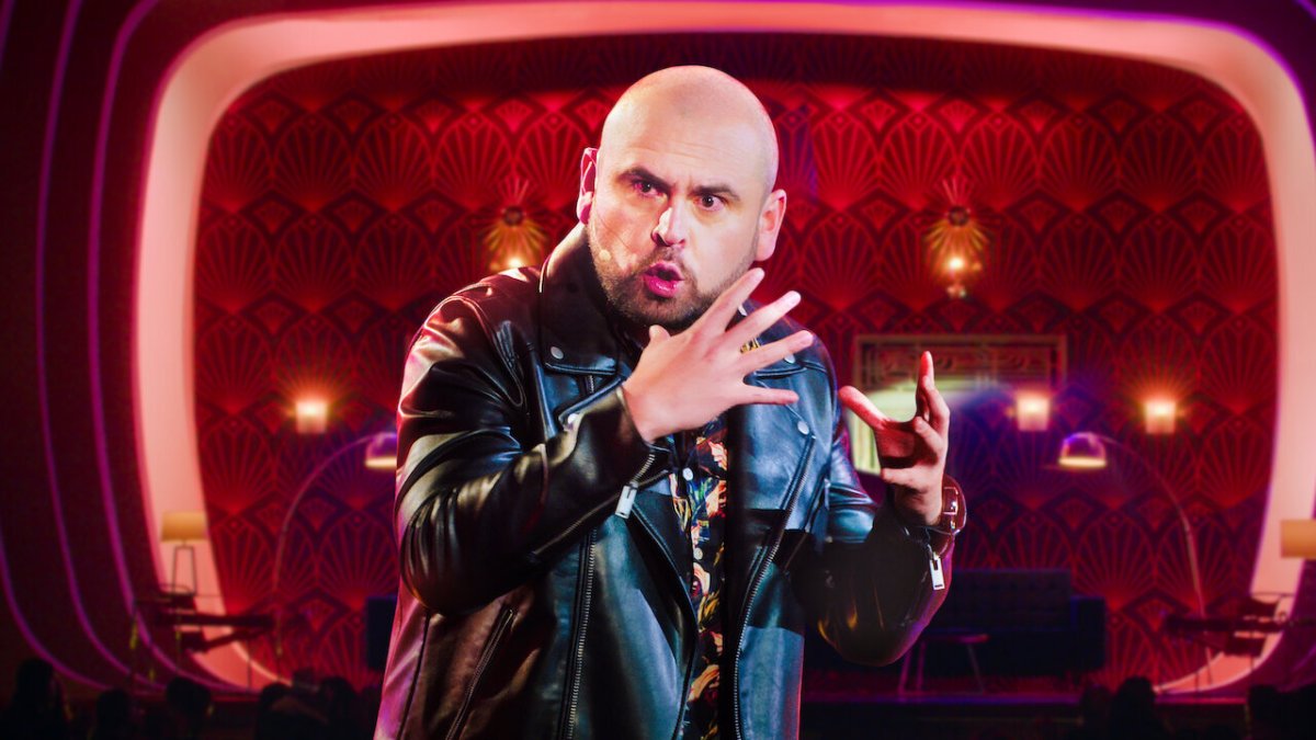 Photograph of comedian Ricardo Quevedo, a bald bearded man in a leather jacket, holding his hands up in front of his face to make a point