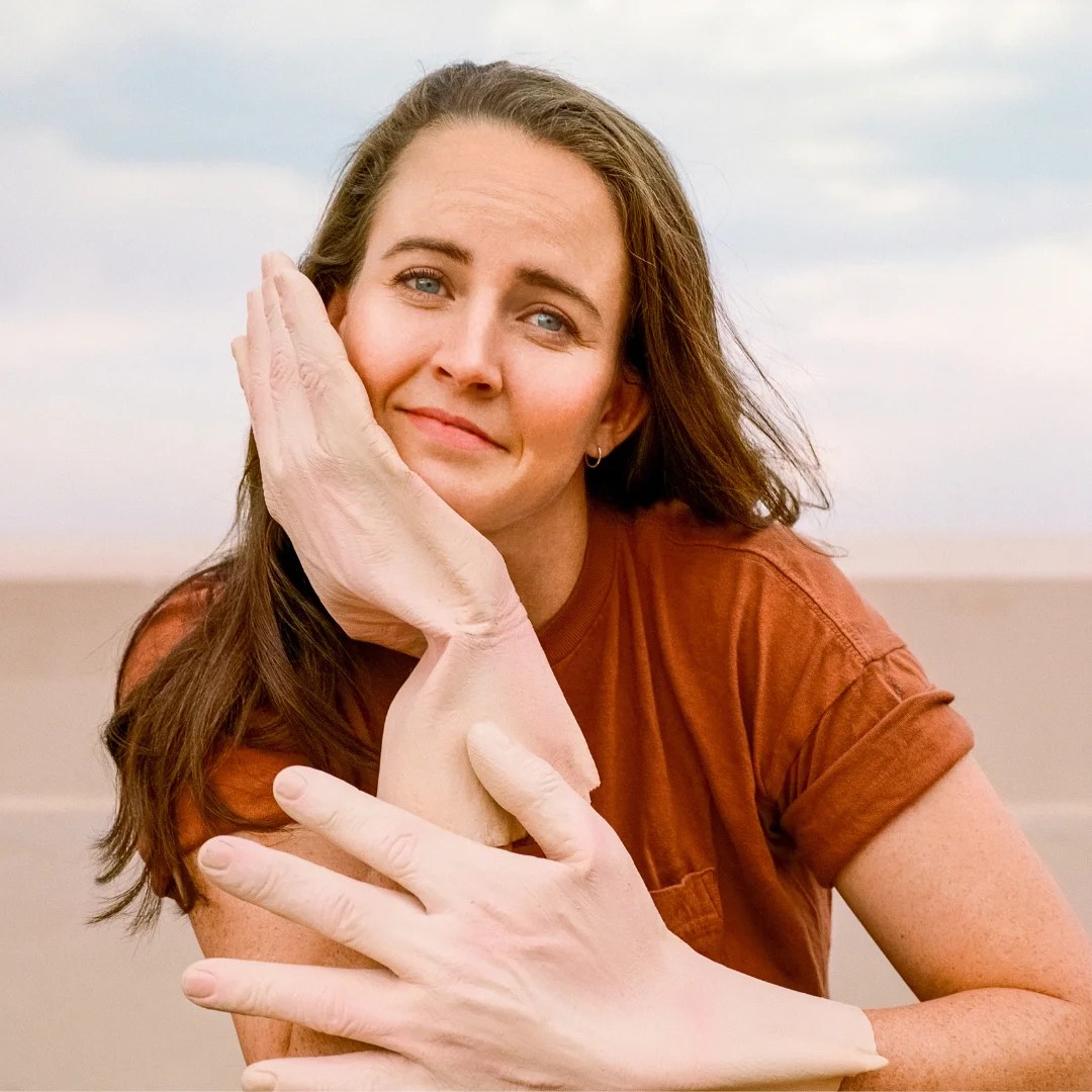 A photograph of comedian Zoë Coombs Marr, a Caucasian woman with long brown hair and a short-sleeved brown T shirt. She is resting her chin on one hand, but both hands are wearing oversize gloves that look like real hands.