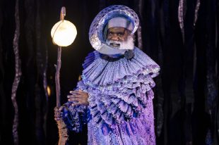 An Aboriginal man in a silver costume has a moon like headdress and carries a lit sphere on a pole. Emu in the Sun, Blak in the Room.