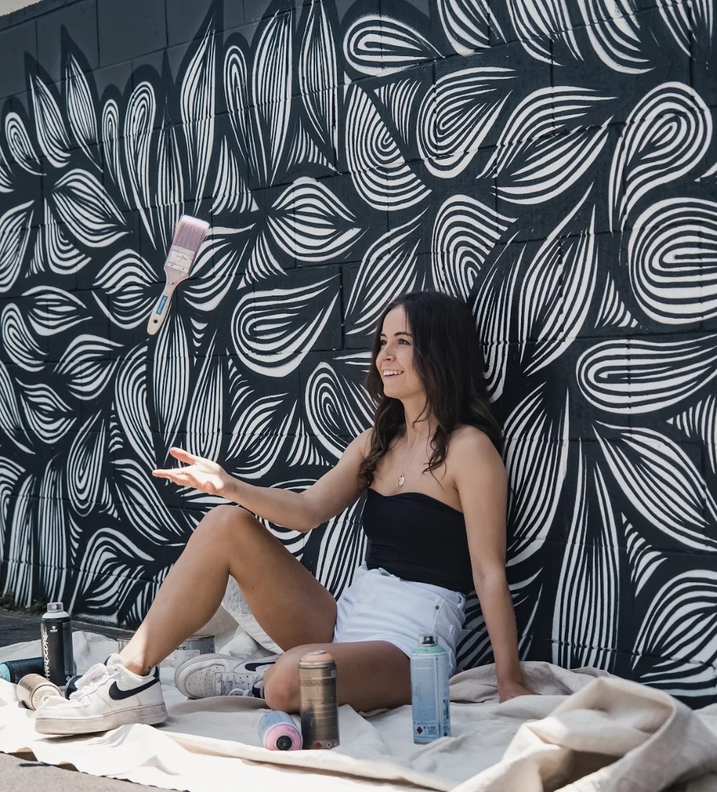 You woman sitting on ground with black and white mural behind, and paint cans around her. Mural artist.