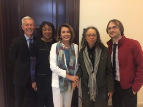 Four Americans of different genders and ethnic backgrounds stand in a line in front of a wooden door, meeting Senator Nancy Pelosi to preserve the National Endowment Agency.