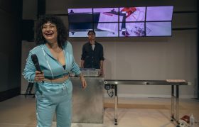 A production of 'Edging.' Sammaneh Pourshafighi in acqua blue tracksuit is in forefront. Eden Falk is in the background. There is an image of the contents of someone's luggage on the screen behind them and part of an airport security equipment with a piece of luggage on it.