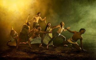 A publicity image for 'Troy', one of seven productions in Malthouse Theatre's 2025 season. A multi-racial cast of seven, including men in skirts and women in tunics reminiscent of ancient Greece, link hands as they struggle across a dramatically lit and smoky battlefield.